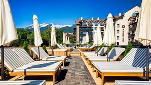 A sunlit outdoor lounge area with striped lounge chairs and umbrellas, a tiled walkway, and a mountain backdrop.
