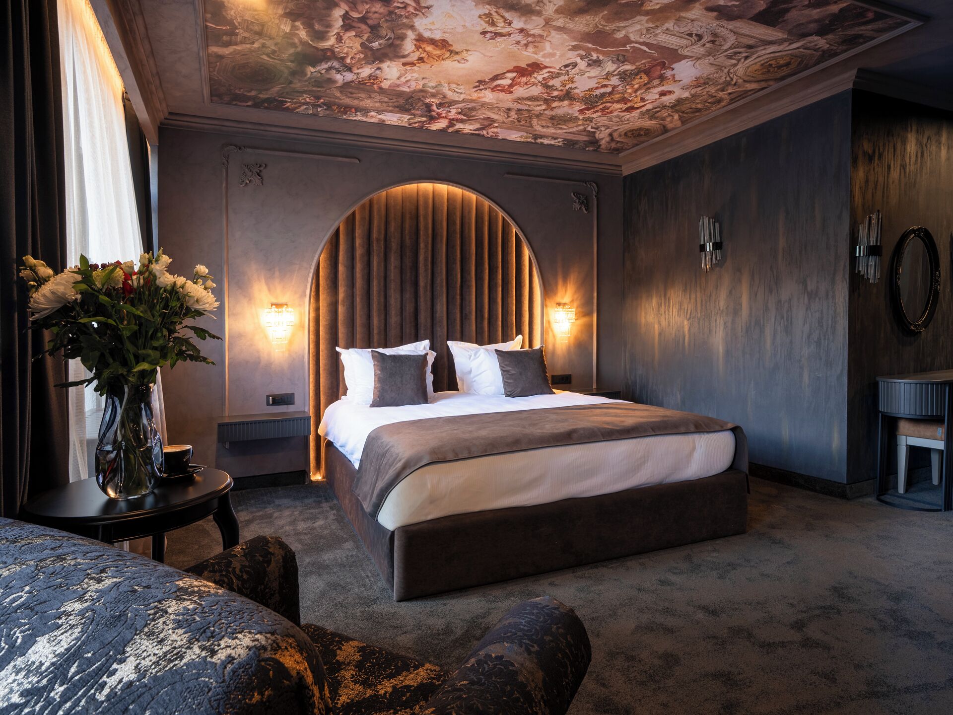 A large bed with a curved headboard, a vase of flowers, and a wall-mounted light fixture.