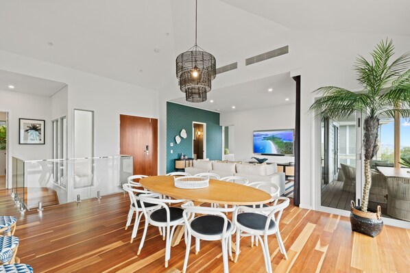 A modern living room with a wooden dining table, white chairs, a large TV, and a large window with a view.