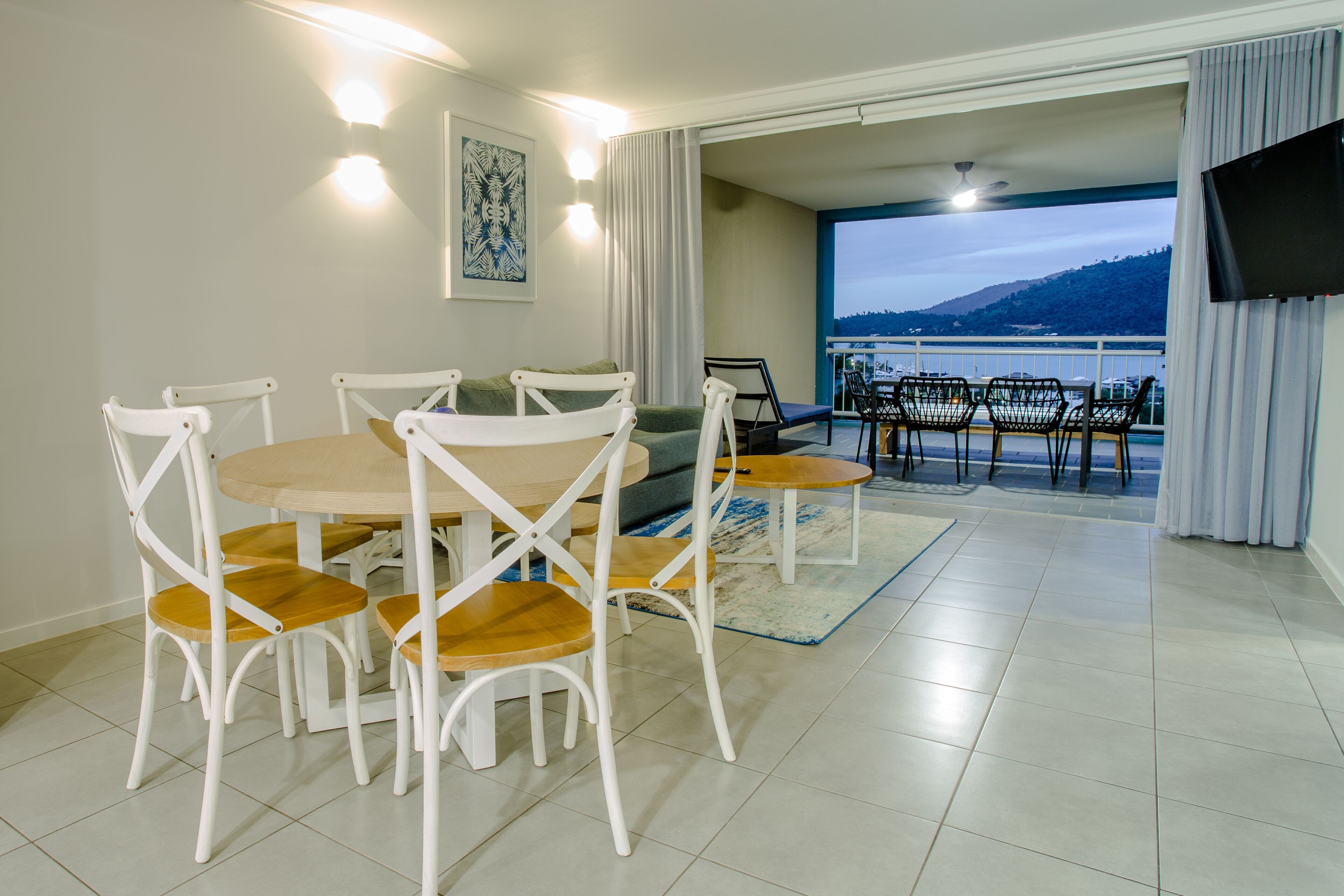 A dining area with a round table and chairs, a sofa, and a view of the sea through a balcony.