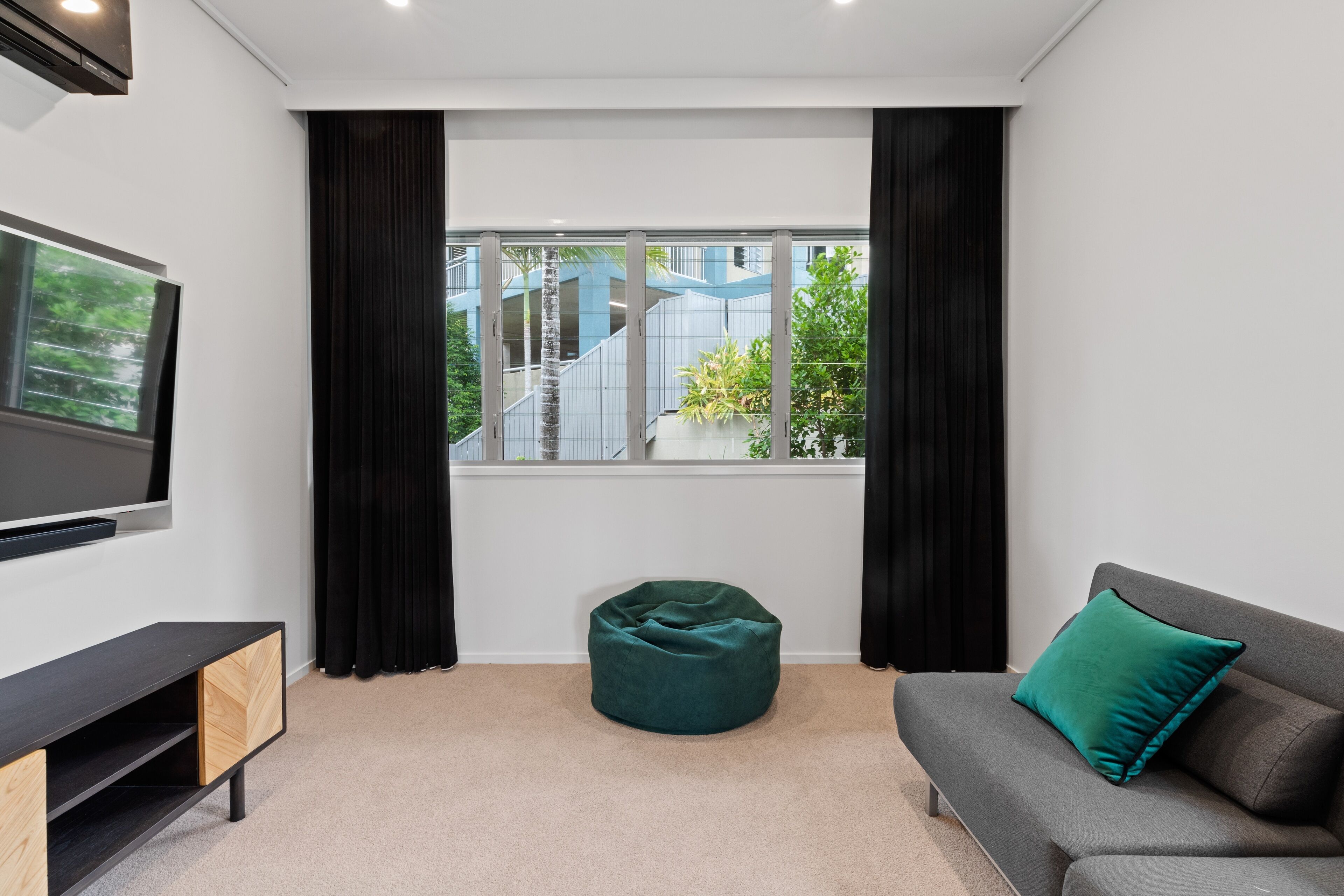 A modern living room with a grey sofa, a teal bean bag, a flat-screen TV, and a window with a view of a building and greenery.