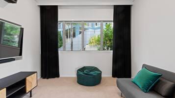 A modern living room with a grey sofa, a teal bean bag, a flat-screen TV, and a window with a view of a building and greenery.