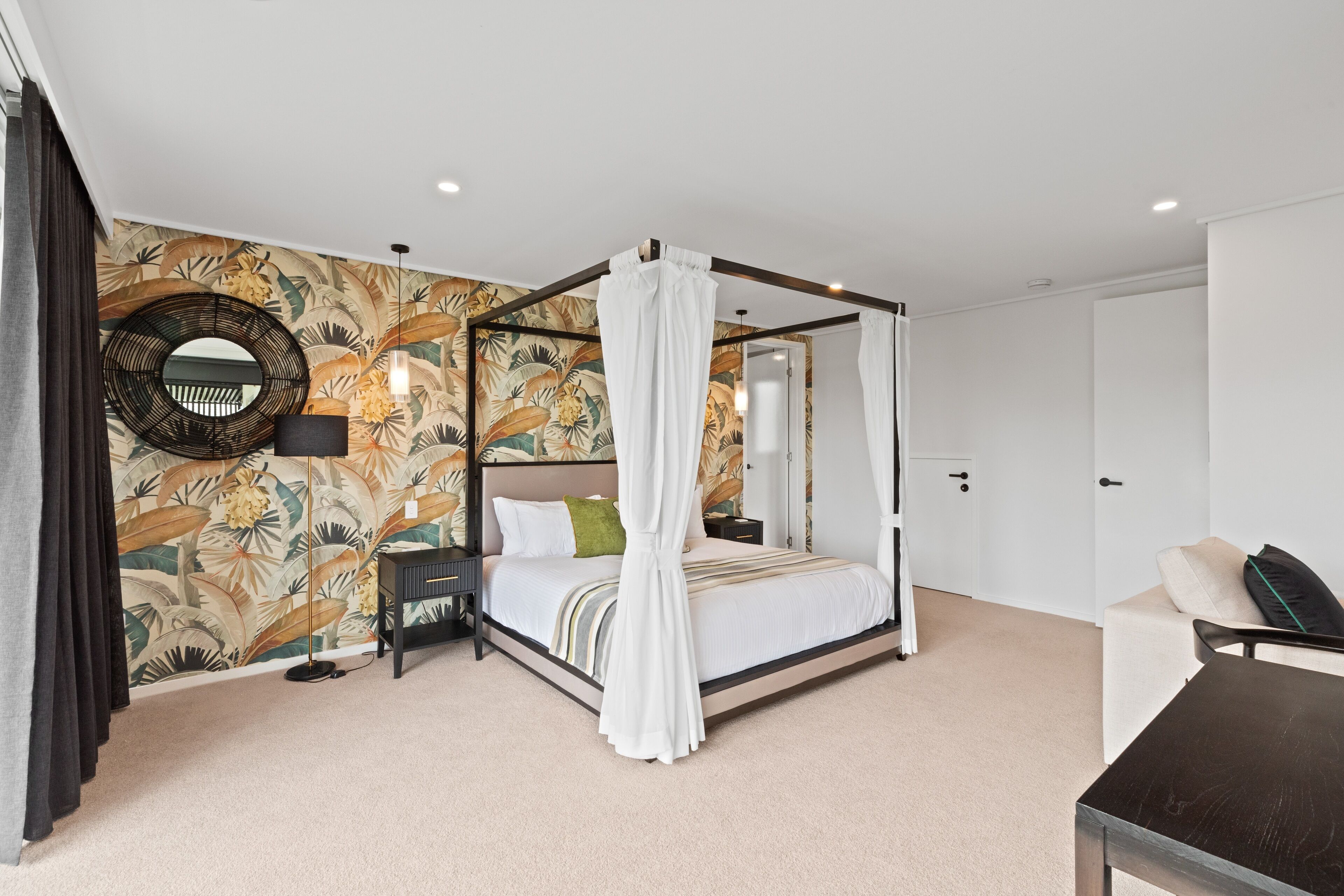 A four-poster bed with a canopy, a round mirror, a nightstand, a lamp, and a sofa in a bedroom with a tropical wallpaper.
