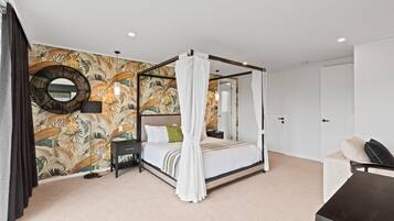 A four-poster bed with a canopy, a round mirror, a nightstand, a lamp, and a sofa in a bedroom with a tropical wallpaper.