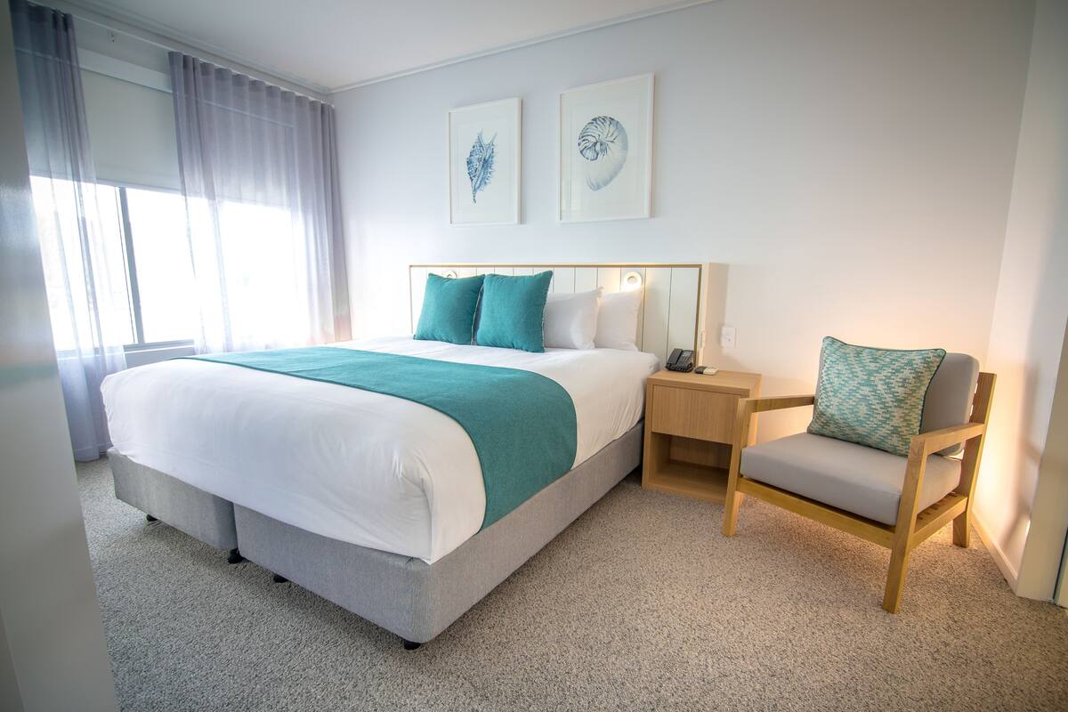 Un dormitorio ordenado con cama, mesita de noche, silla y una obra de arte en la pared.