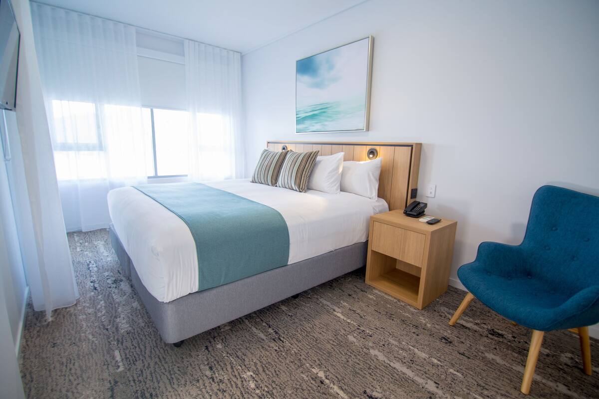 Habitación de hotel con una cama grande, una mesita de noche con teléfono, una silla azul y un cuadro en la pared.