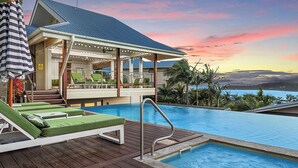 An infinity pool with lounge chairs, a covered seating area, and a view of the ocean at sunset.