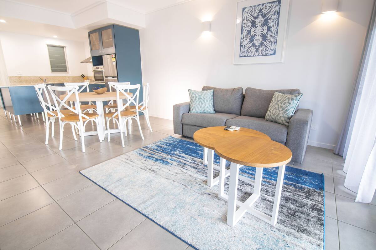 Una sala moderna con un sofá gris, una mesa de centro de madera, y un comedor con sillas blancas y una isla azul.