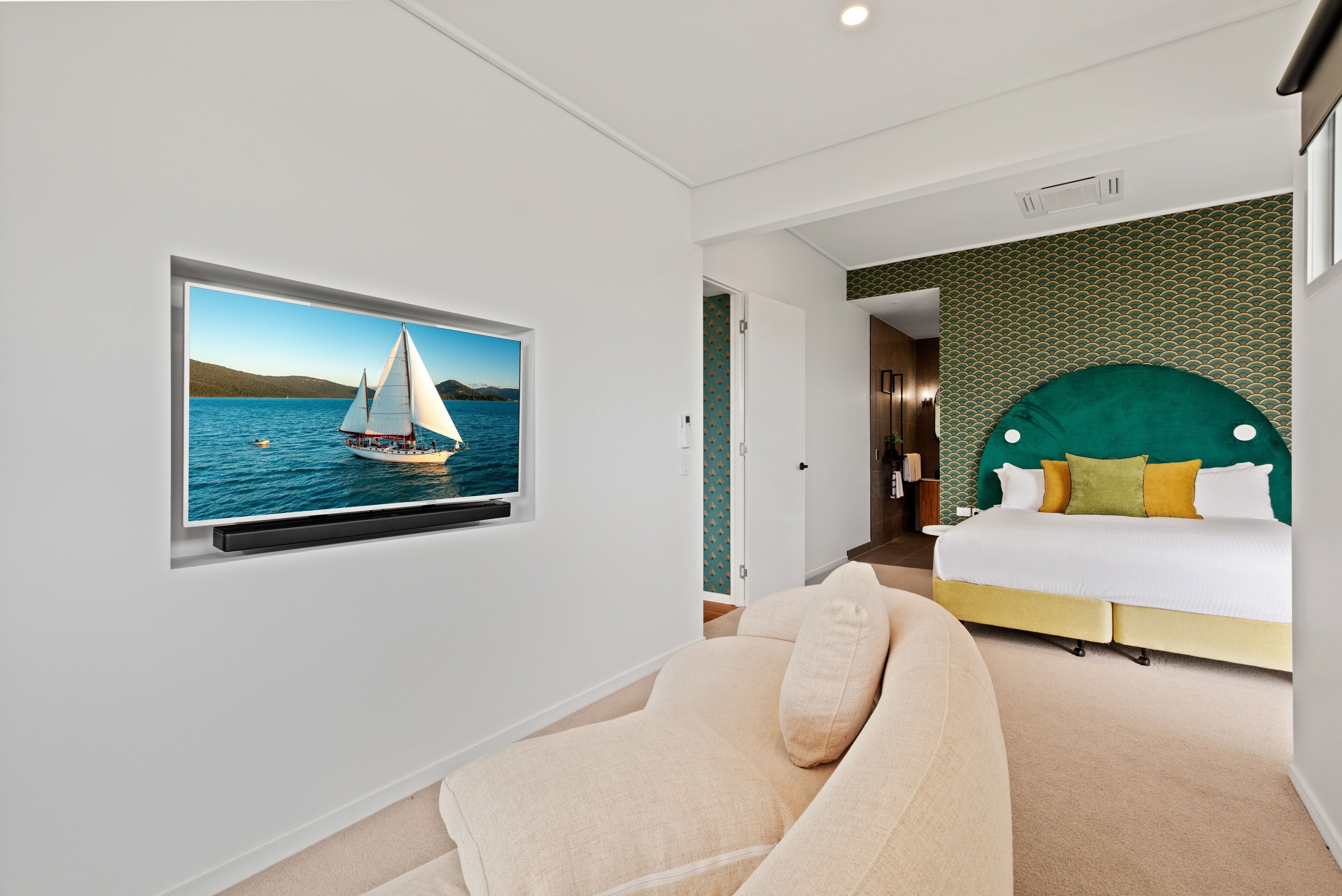 A modern hotel room with a large TV displaying a sailboat on water, a bed with a green headboard, and a beige sofa.