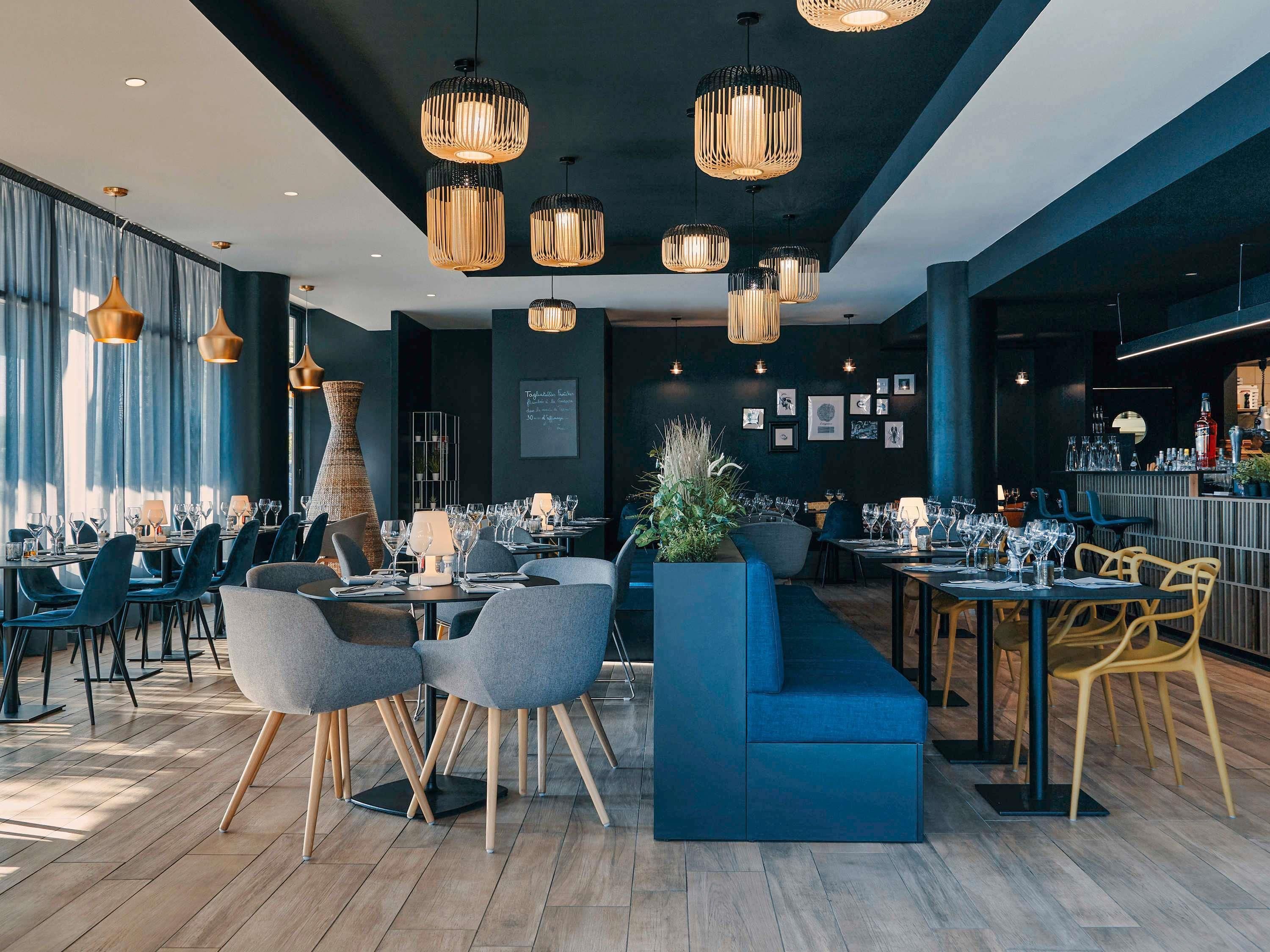 Interior moderno de restaurante con techos altos, iluminación colgante y una variedad de arreglos de asientos, incluyendo mesas y un sofá azul.