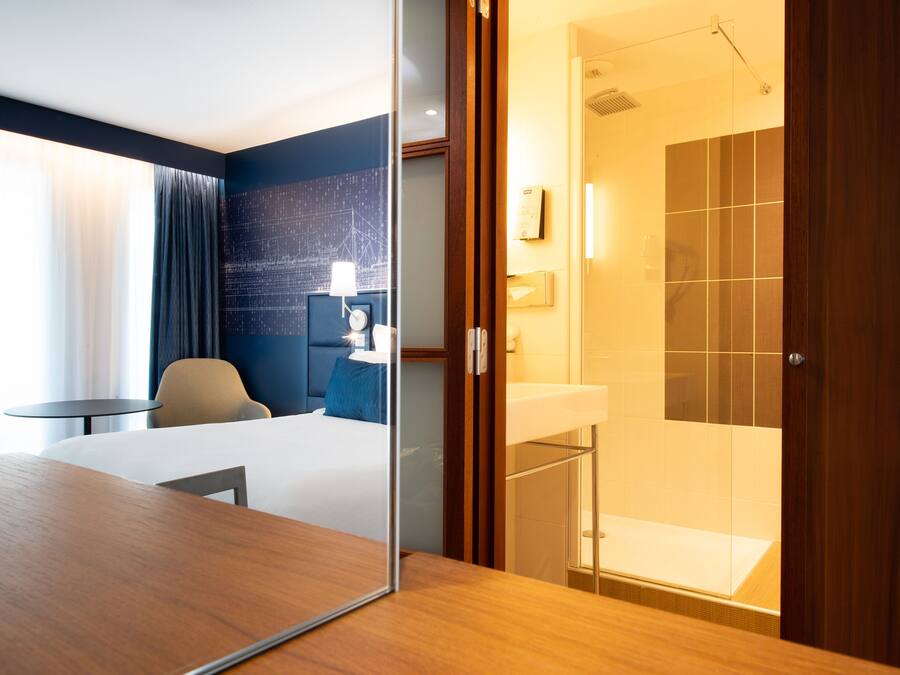 Hotelkamer met een houten bureau, een bed, een stoel, een klein tafeltje, een glazen scheidingswand en een badkamer met douche, plus een muur met een geometrisch patroon.