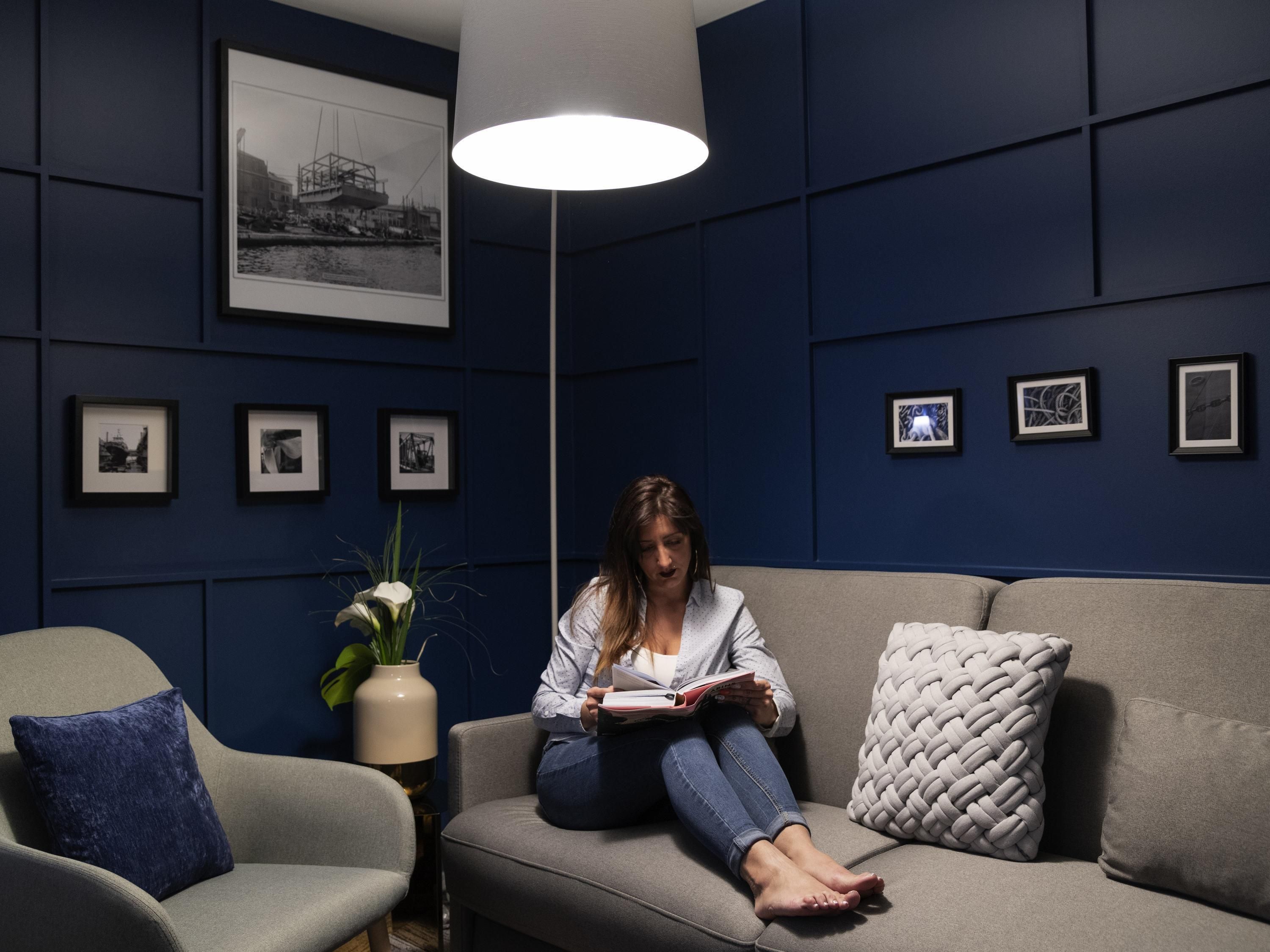 Una persona sentada en un sofá en una habitación con pared azul oscuro, leyendo un libro. Hay cuadros en la pared y una lámpara de pie.