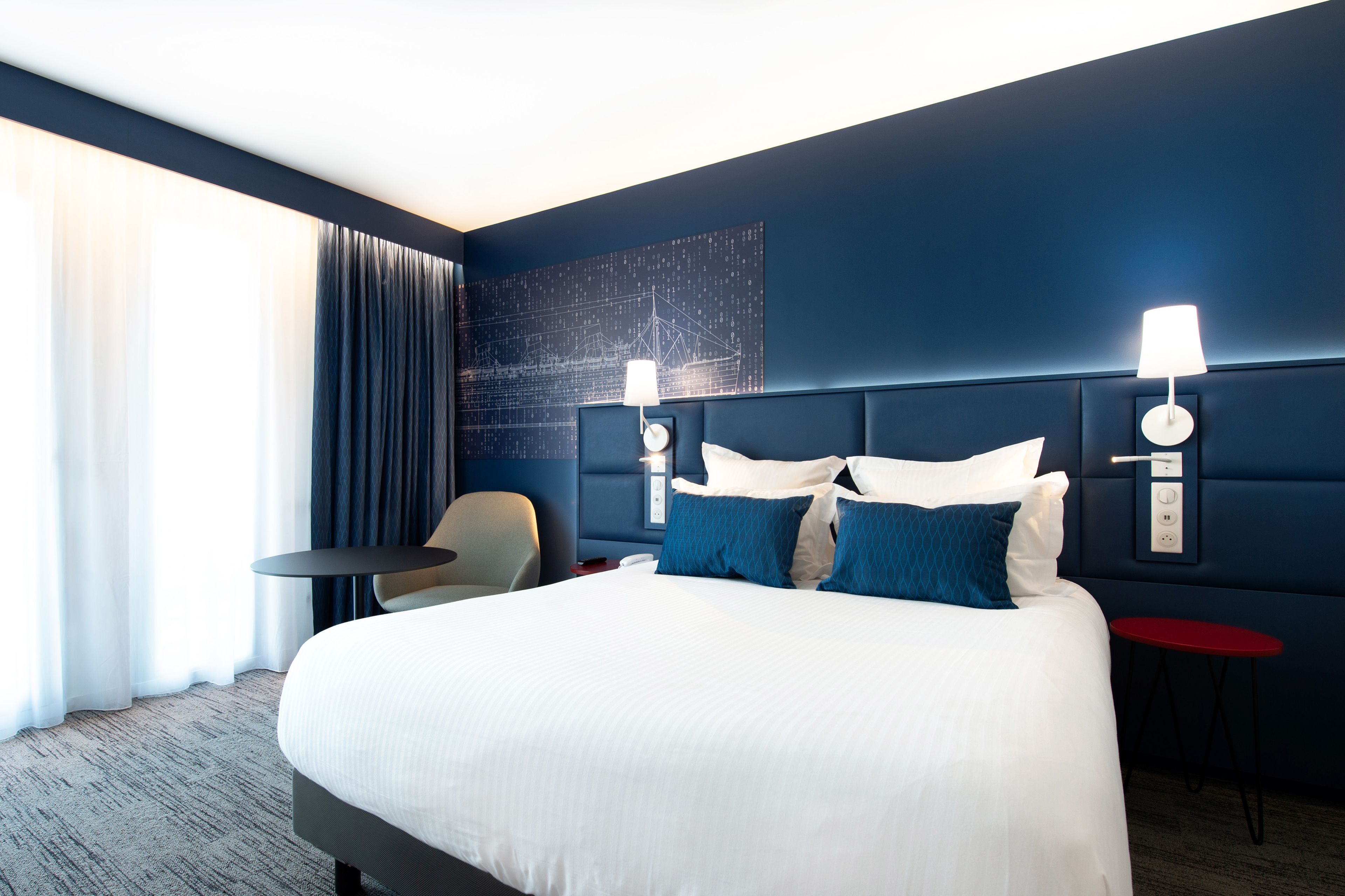 Ein Hotelzimmer mit einem großen Bett, zwei Nachttischen, einem Sessel und einem roten Beistelltisch, sowie einer Wandlampe.