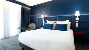 A hotel room with a large bed, two bedside tables, a chair, a red side table, and a wall-mounted lamp. - Hôtel Mercure Toulon La Seyne-Sur-Mer (La Seyne-sur-Mer)