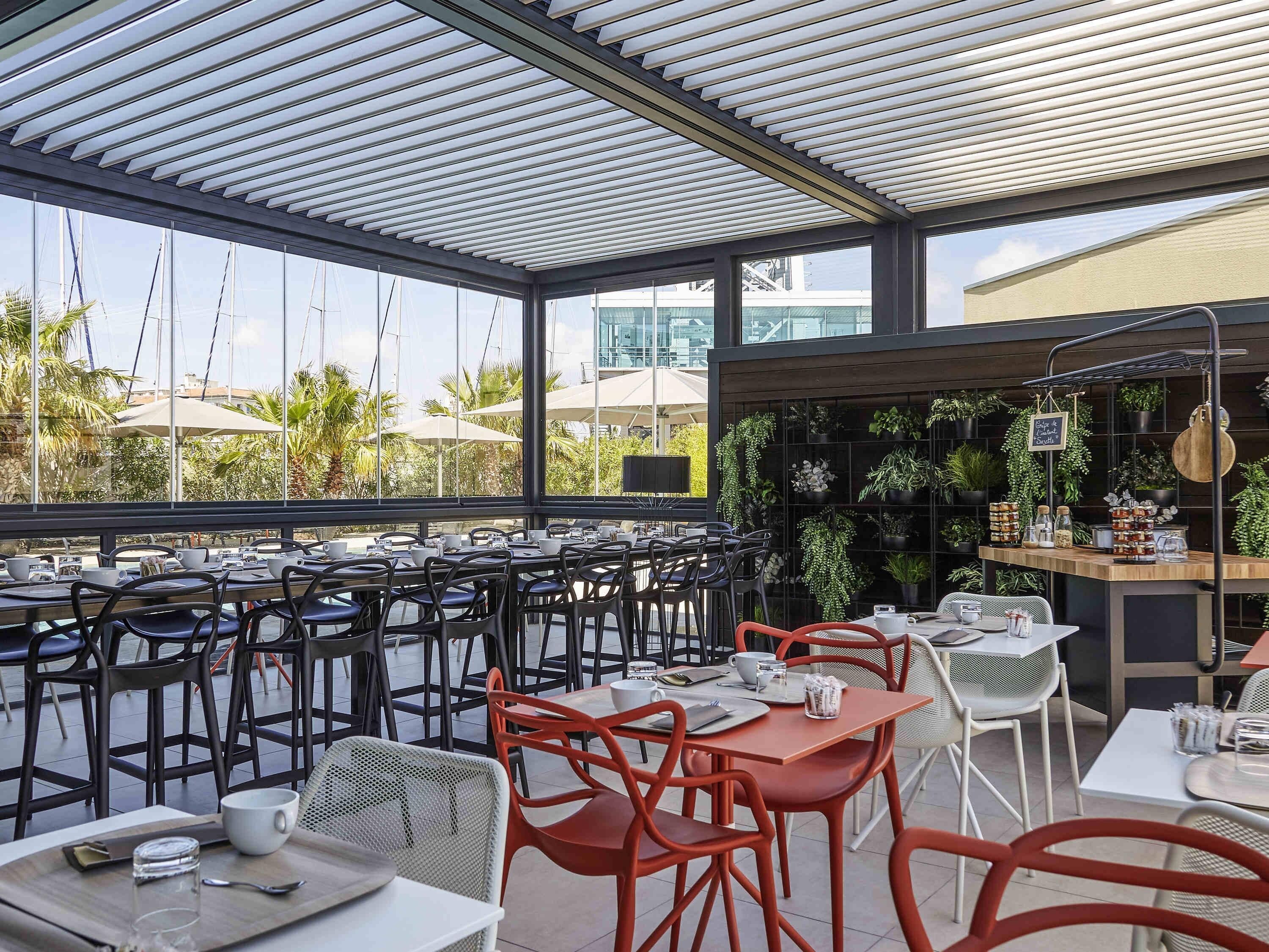 Un área de comedor al aire libre con mesas y sillas, una pared de plantas en macetas, y vista al mar y edificios al fondo.