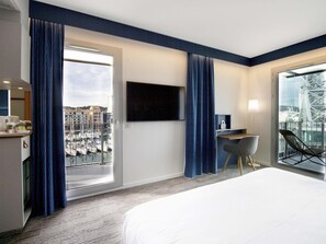 Habitación de hotel con cama, escritorio, televisor y balcón con vista a barcos y edificios.