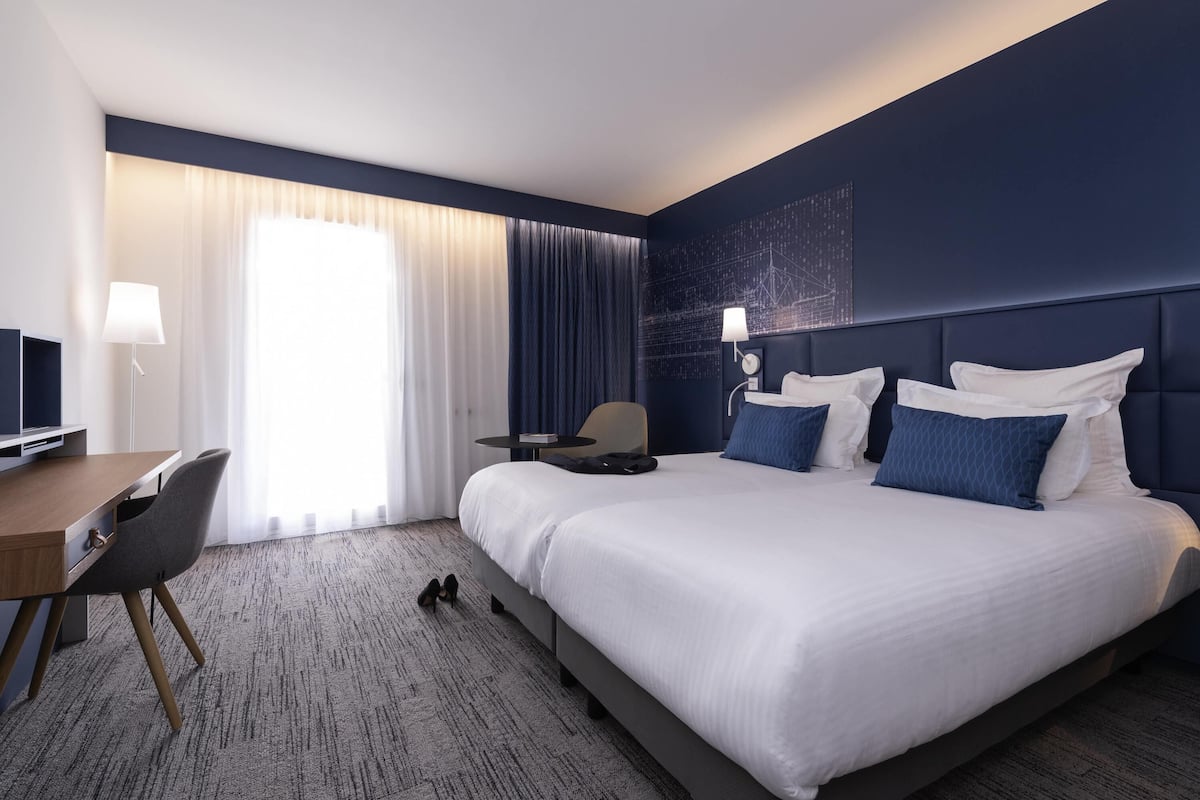 Une chambre d’hôtel avec un grand lit, un bureau avec une chaise, une télévision et une fenêtre avec des rideaux.