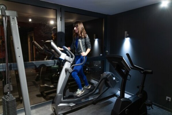 Una persona usando una bicicleta estática en una habitación con un ventanal grande y una zona de equipo de gimnasio visible.