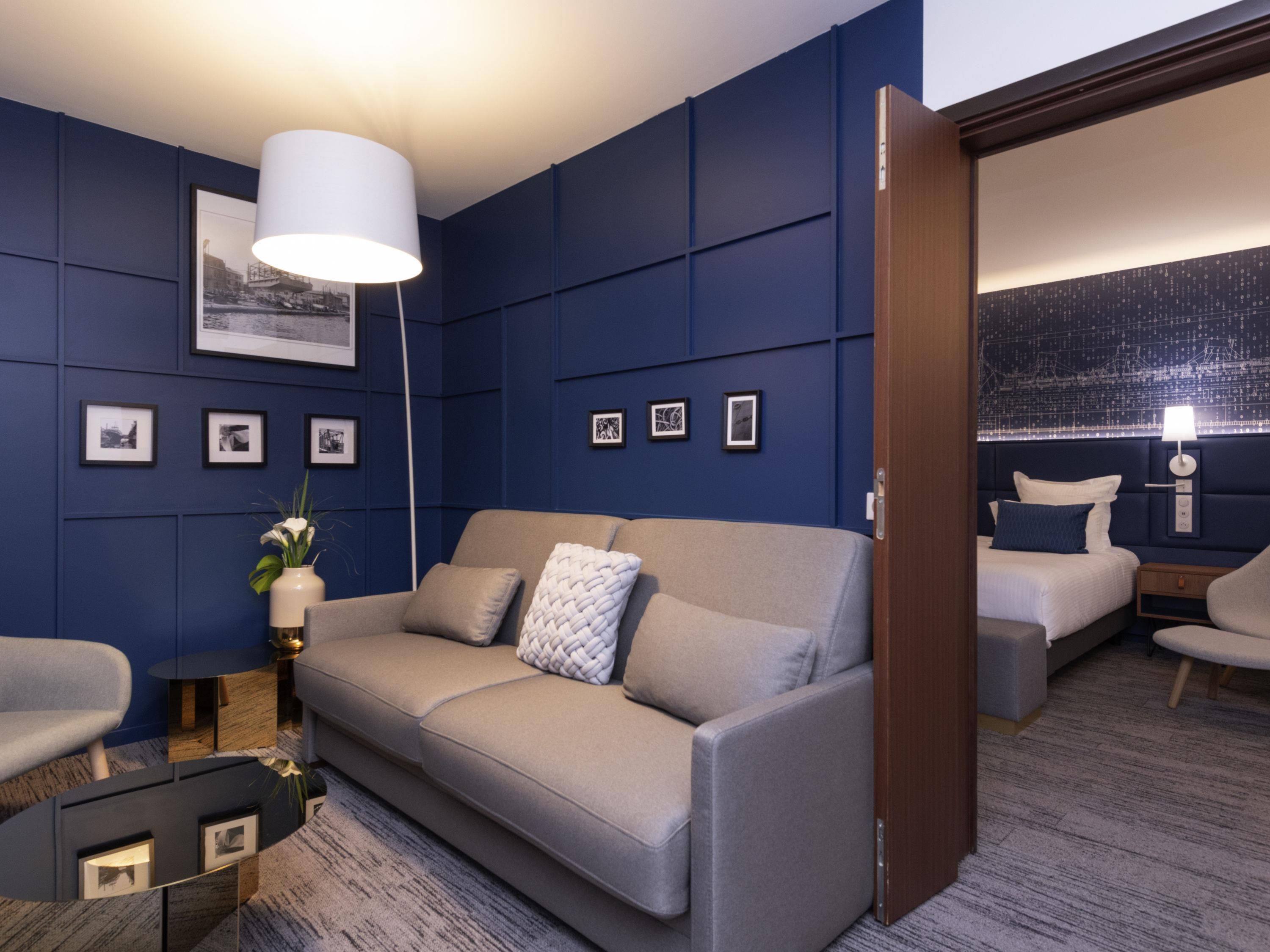 Ein modernes Hotelzimmer mit einem grauen Sofa, einem Beistelltisch mit einer Vase, gerahmten Bildern an der Wand und einem großen Fenster mit Blick.