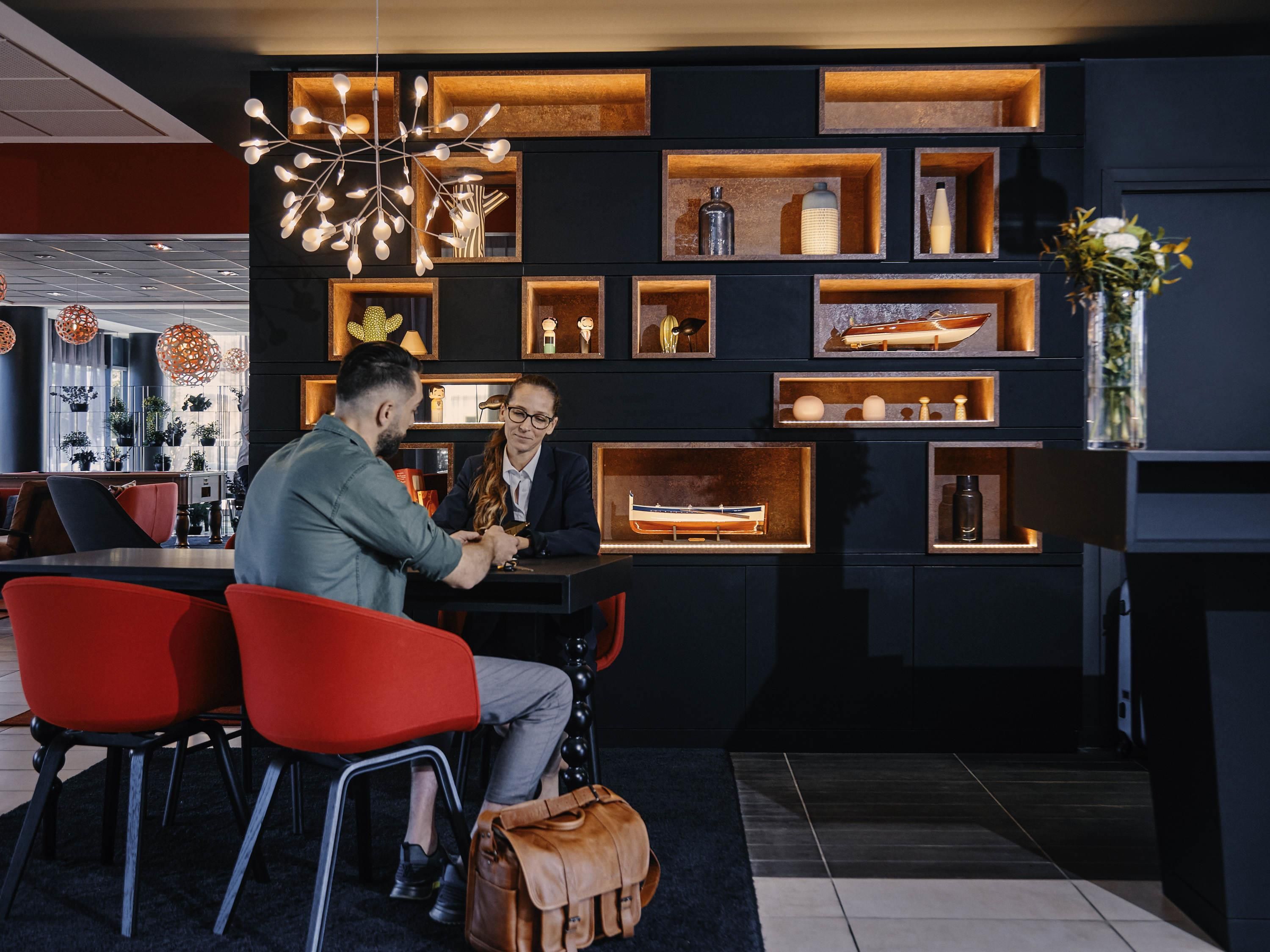 Un interior moderno con una paleta de colores oscuros, que presenta una zona de estar con sillas rojas, una mesa y una pared decorativa con estanterías que exhiben diversos objetos.