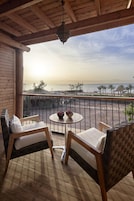 A wooden balcony with two wicker chairs, a small round table with a fruit bowl, and a view of the sea and palm trees.