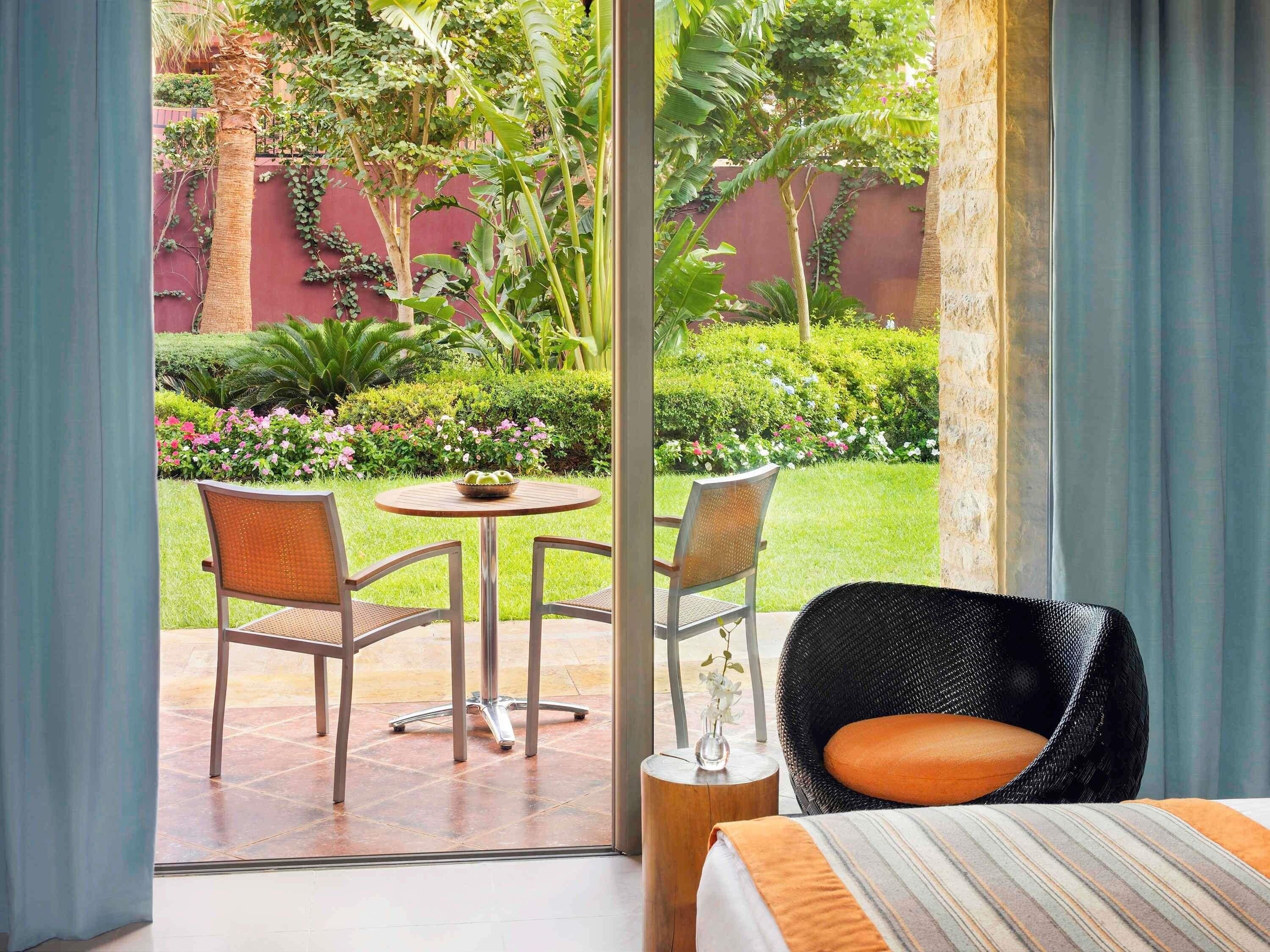 A hotel room with a large sliding glass door leading to an outdoor patio with two wicker chairs, a small round table, and a bed with a striped bedspread.