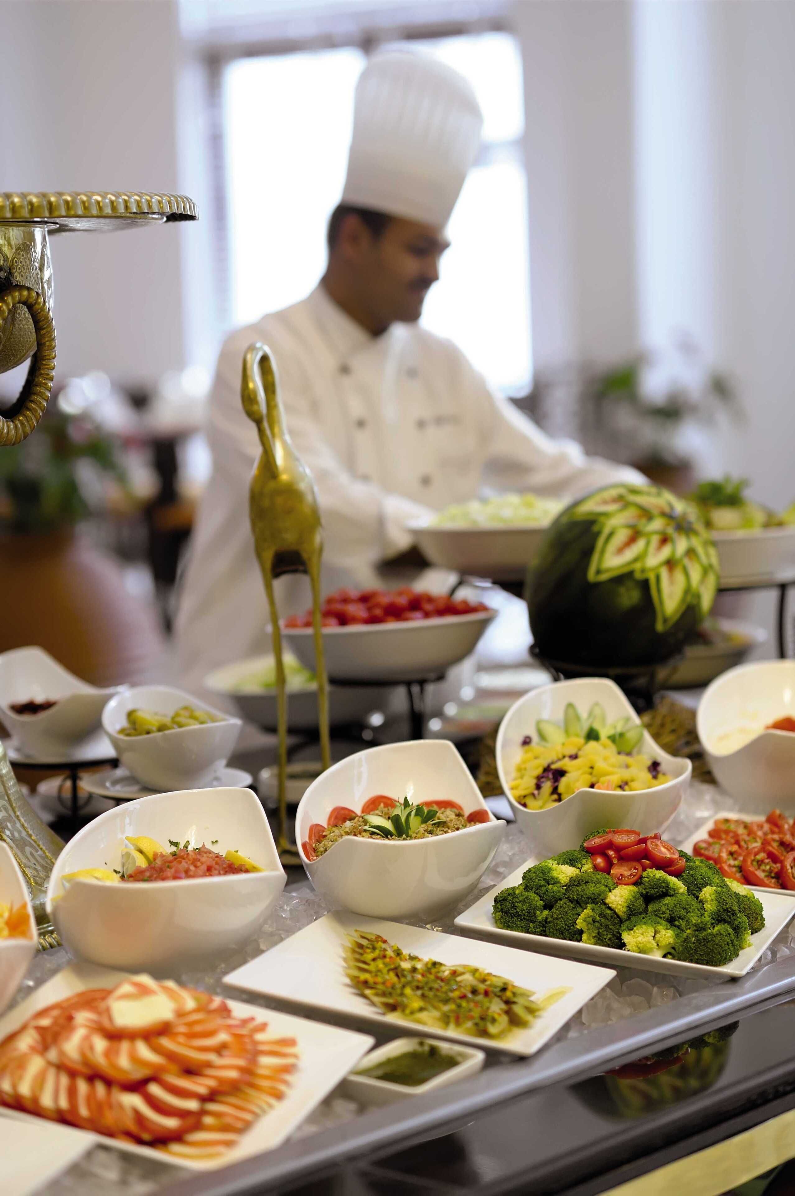 Un buffet proposant divers plats, dont une pastèque sculptée en forme décorative, et un chef cuisinier en arrière-plan.