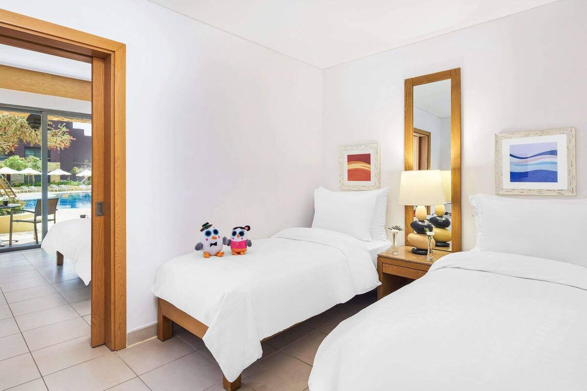 A hotel room with two beds, a large window, a wooden door, and a view of a swimming pool.