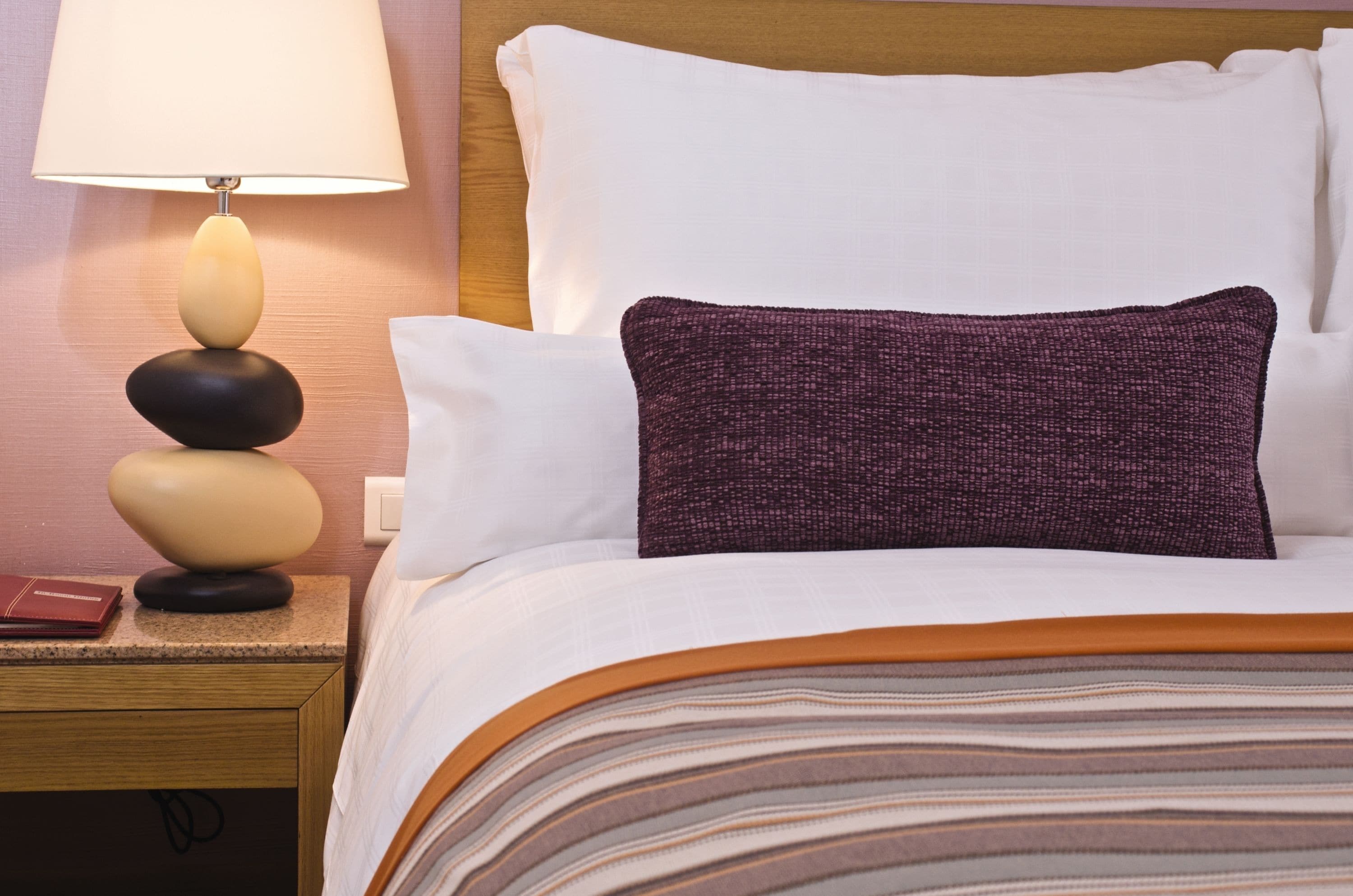Una cama bien hecha con una colcha a rayas, una lámpara con pantalla color beige y una almohada oscura.