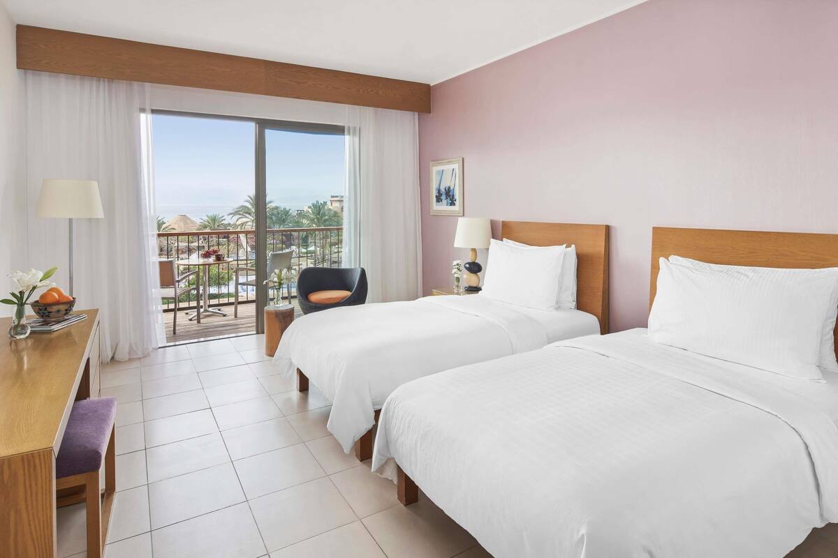 A hotel room with two beds, a desk, a chair, and a balcony with a view of the ocean and palm trees.