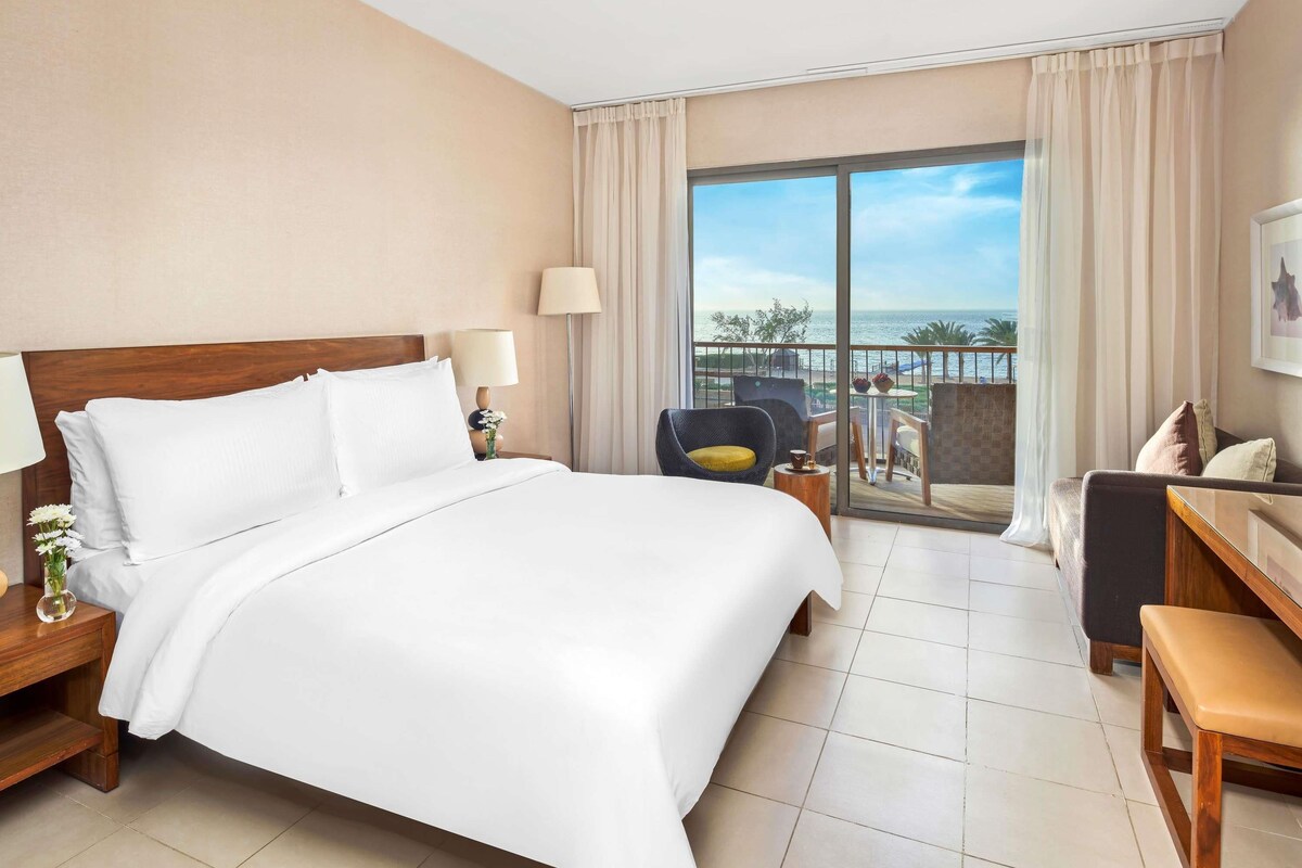 A hotel room with a large bed, a desk, a chair, and a balcony with a view of the sea.