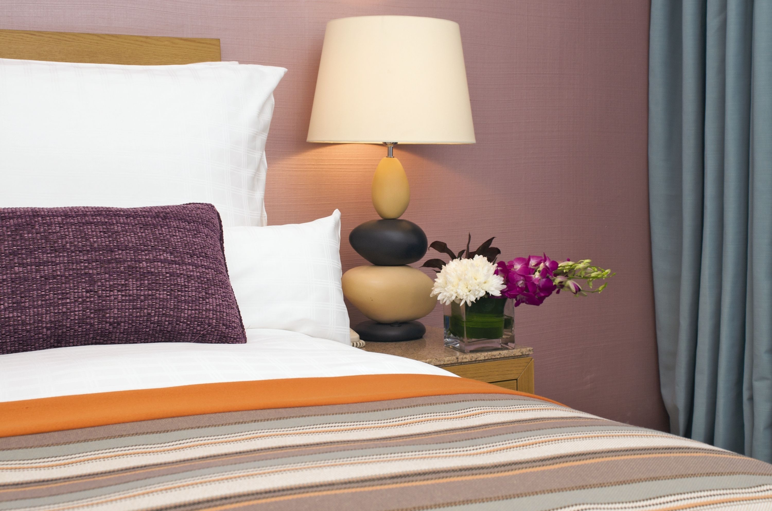 Una cama bien hecha con una colcha a rayas, una almohada blanca, un cojín morado y una mesita de noche con una lámpara y un florero con flores.