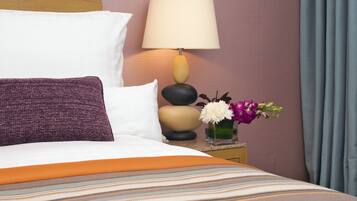 Una cama bien hecha con una colcha a rayas, una almohada blanca, un cojín morado y una mesita de noche con una lámpara y un florero con flores.