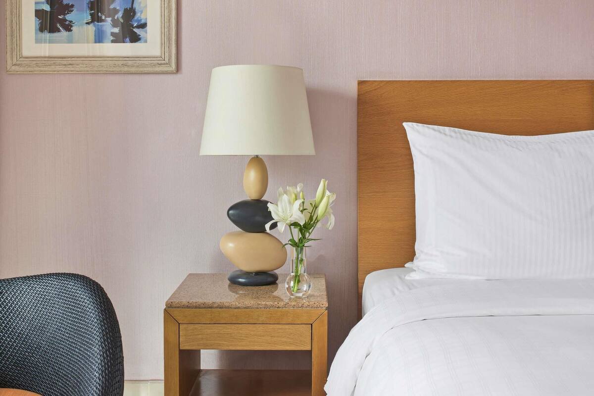 A hotel room with a bed, a nightstand with a lamp and vase, and a chair.