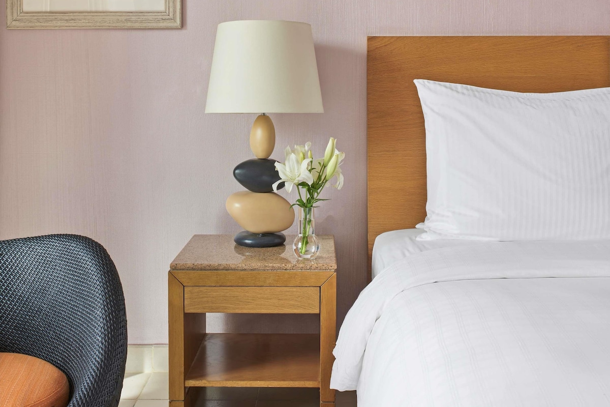 A hotel room with a bed, a nightstand with a lamp and vase, and a chair.