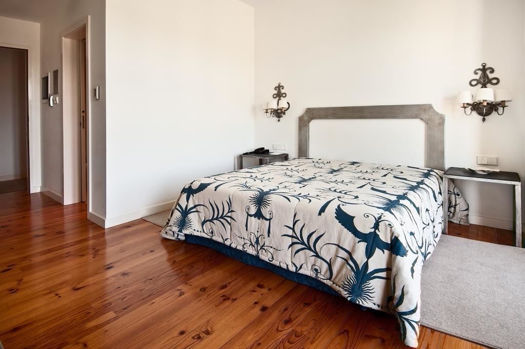 A bedroom with a wooden floor, a bed with a floral bedspread, bedside tables with lamps, and a nightstand.