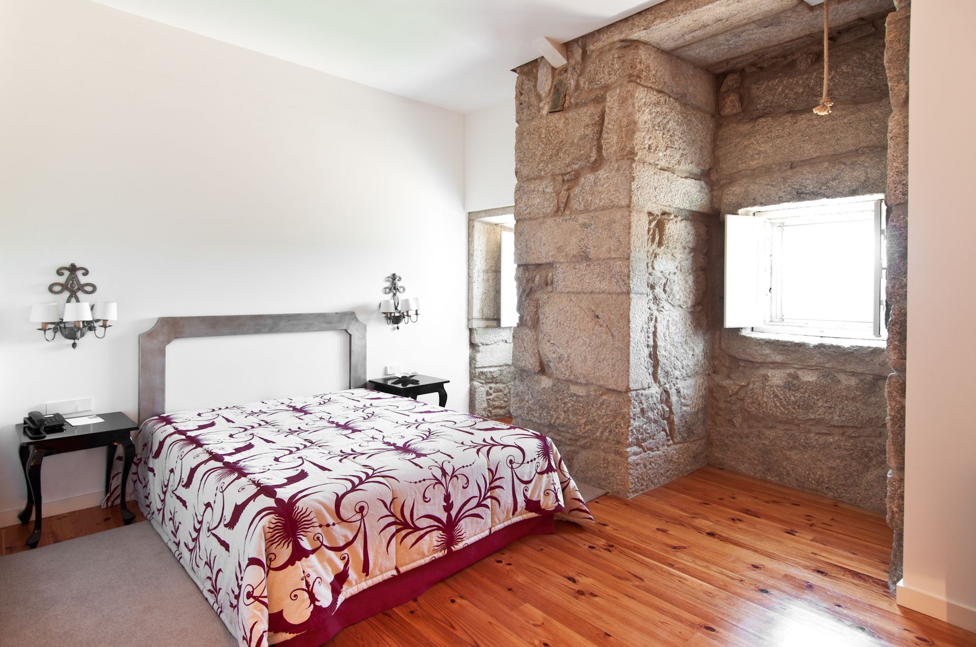 A bedroom with a bed, wooden floor, stone wall, and a window.