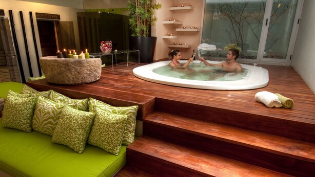 A modern spa area with a wooden deck, a green sofa with patterned cushions, and a hot tub with two people. - Convento dos Capuchos (Monção)