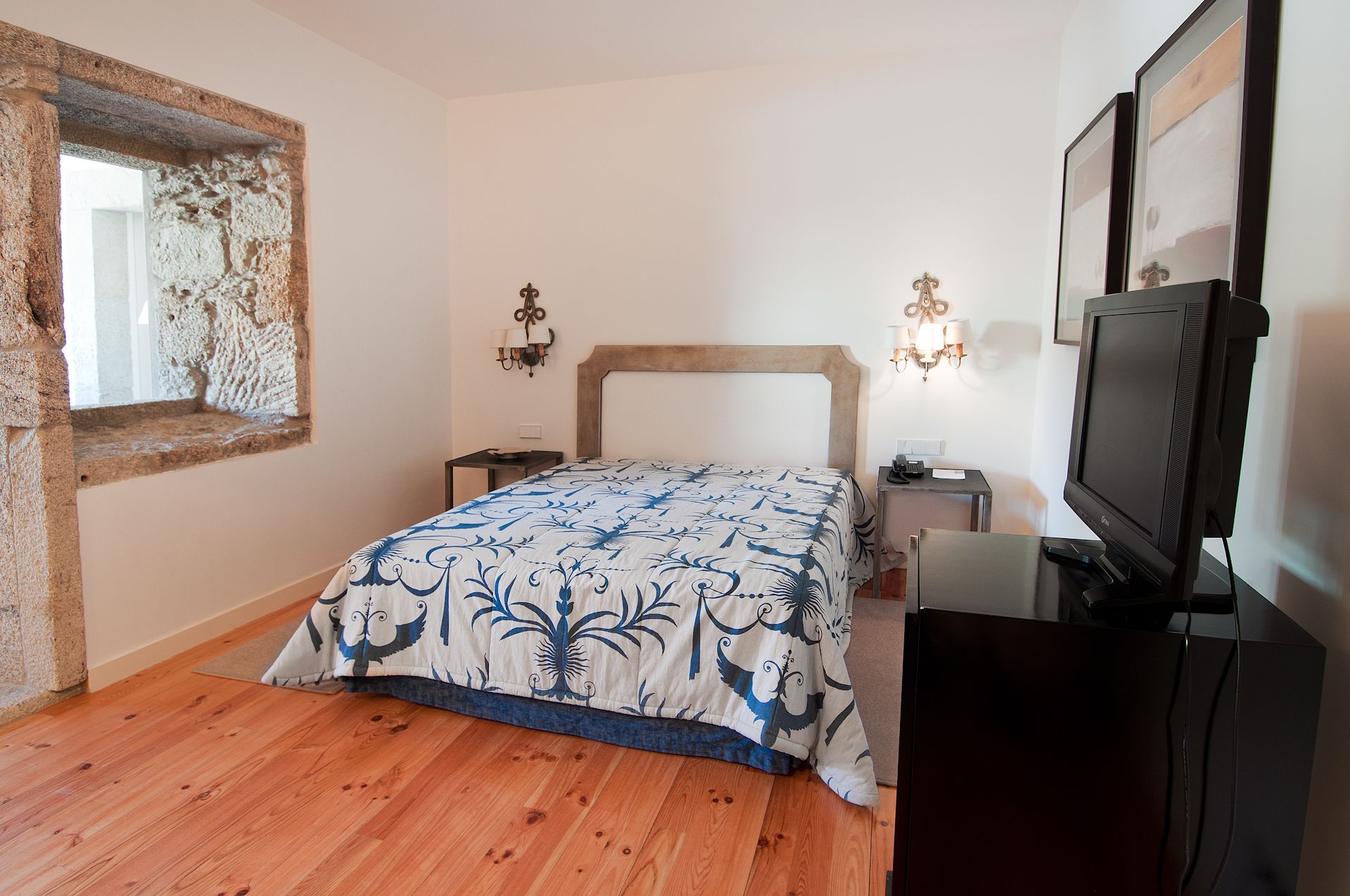 A bedroom with a wooden floor, a bed with a patterned bedspread, a television, and a stone wall.