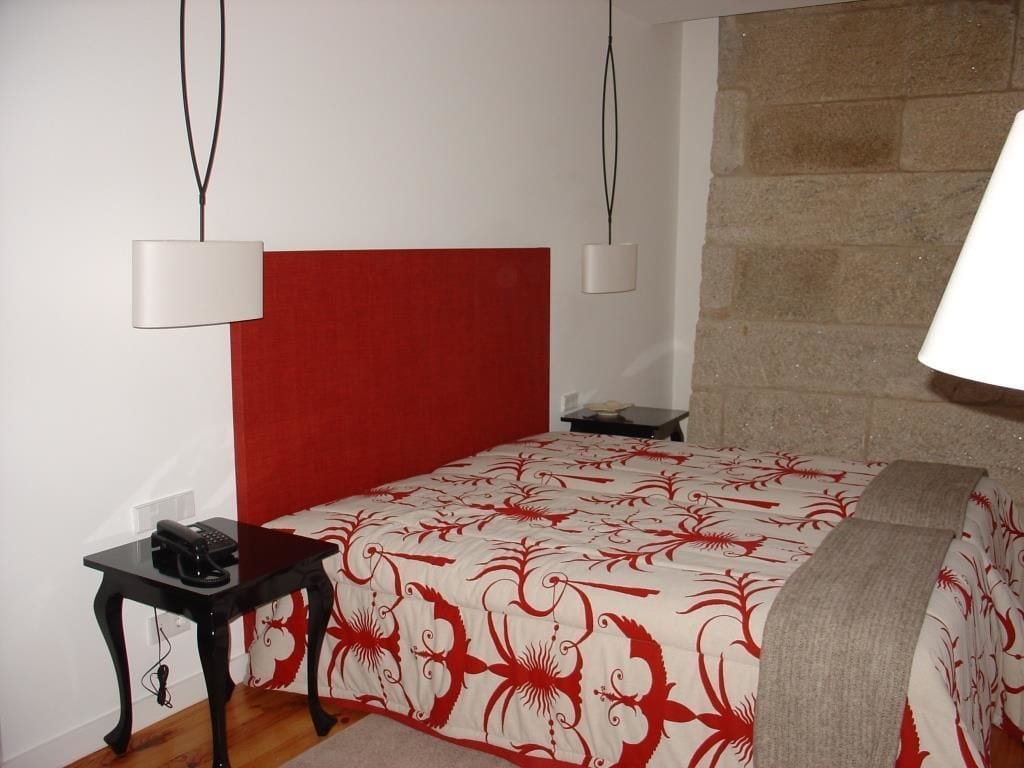 Een slaapkamer met een rode hoofdeinde, een bed met een bloemenprintdeken, een nachtkastje met een lamp en een klein tafeltje met een telefoon.
