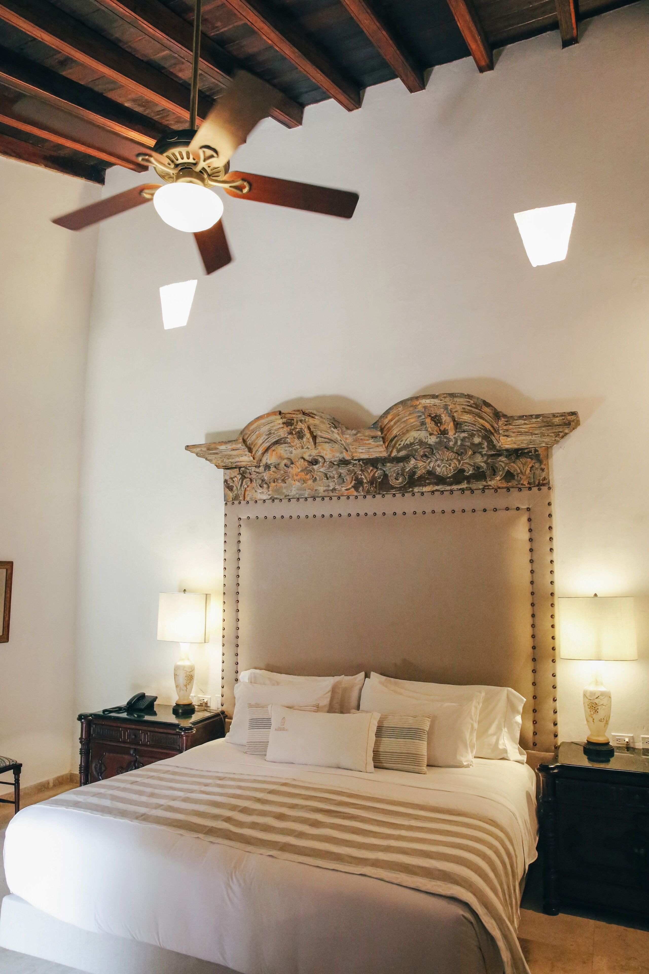 A bedroom with a large bed, a ceiling fan, two bedside lamps, and a decorative headboard.