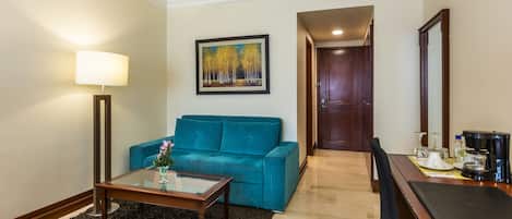 A hotel room with a teal sofa, a glass coffee table, a lamp, a painting on the wall, and a wooden door.
