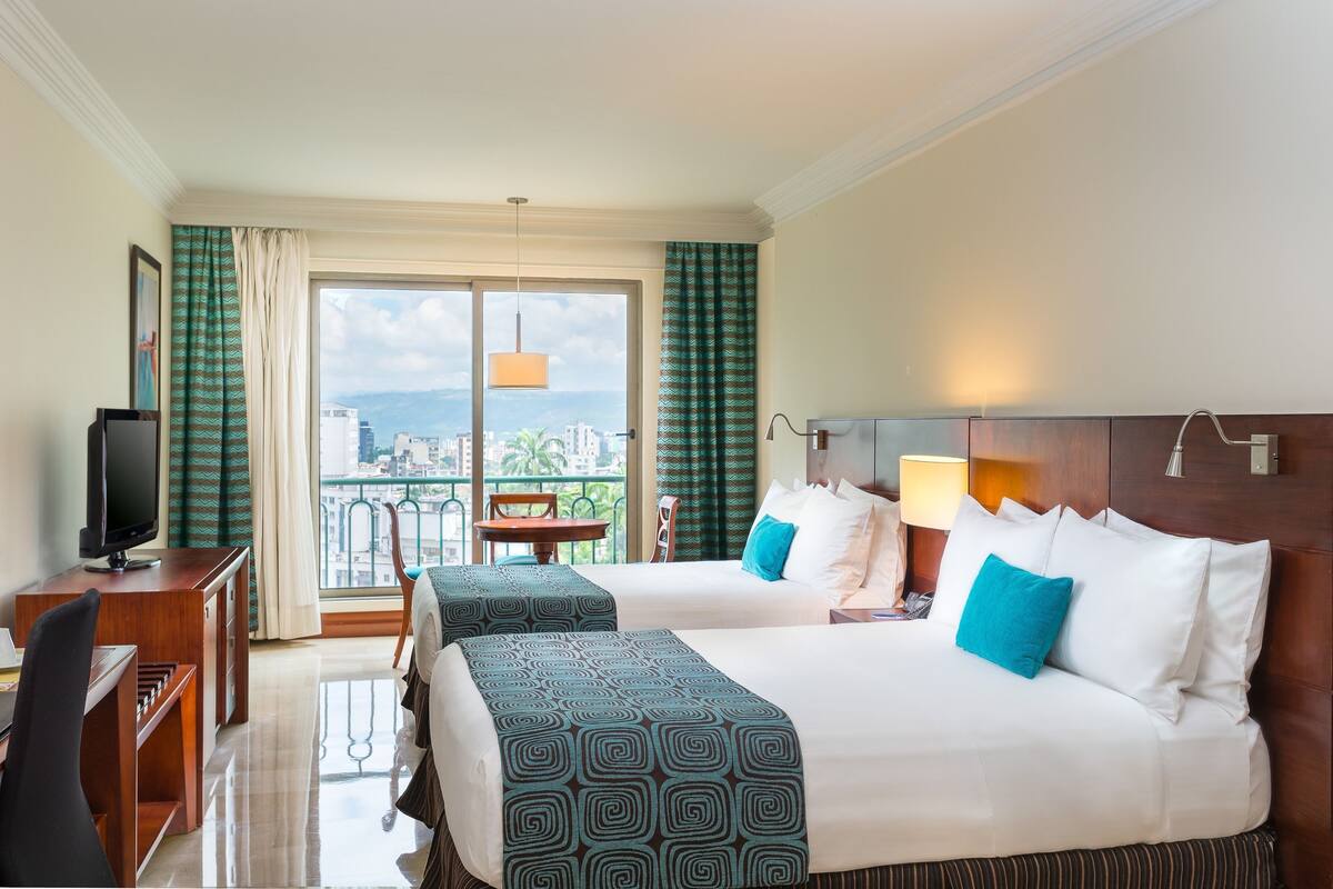 Ein Hotelzimmer mit zwei Betten, einem Schreibtisch, einem Fernseher und einem Balkon mit Blick auf die Stadt.