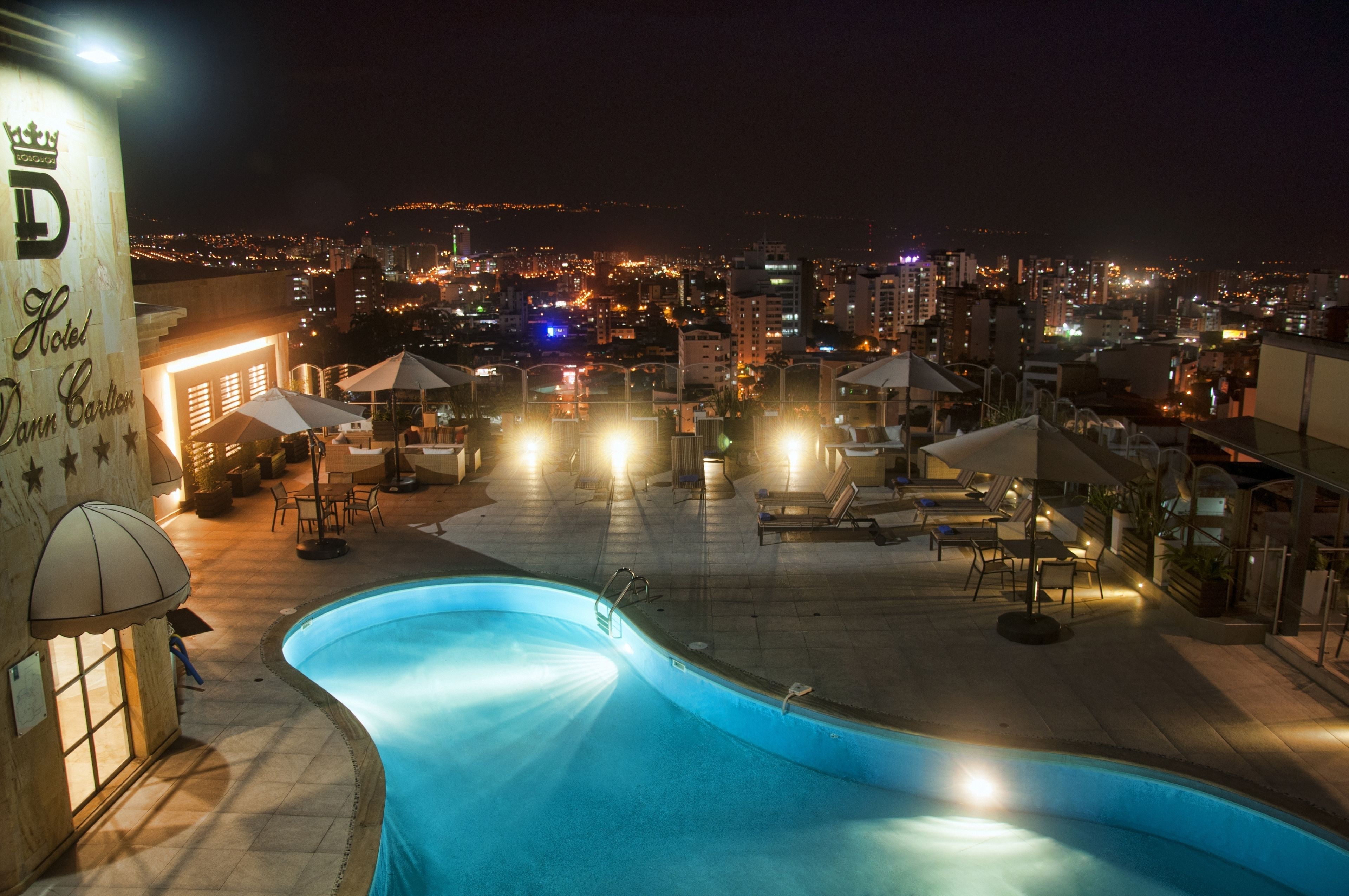 Une piscine située sur le toit d’un hôtel, avec des chaises longues et des parasols, entourée des lumières de la ville la nuit.