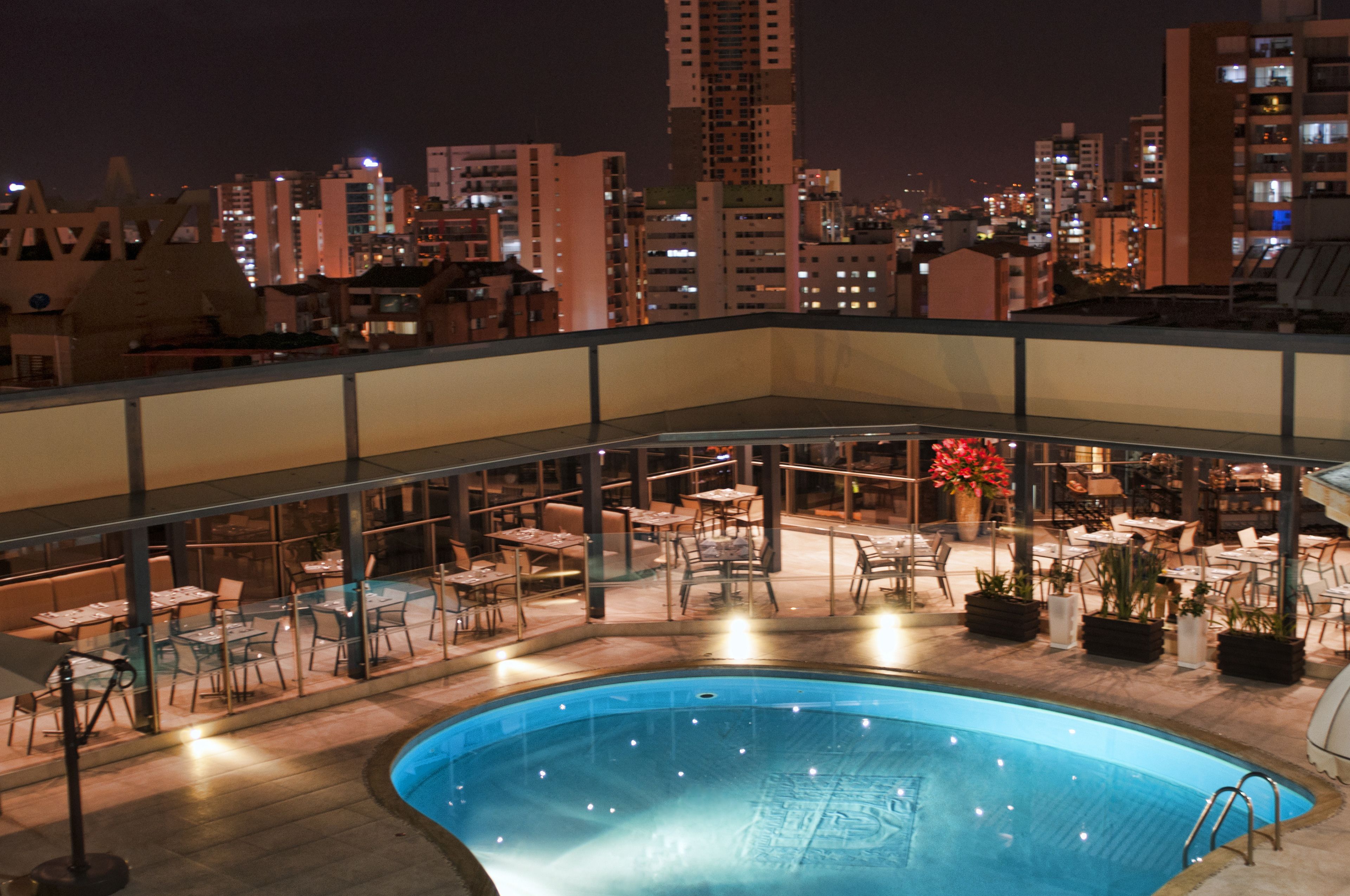 A rooftop pool area with tables and chairs, surrounded by city buildings at night.
