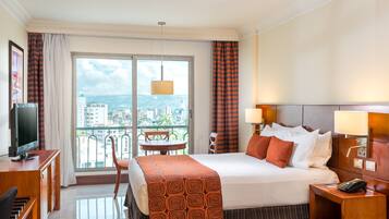 A hotel room with a bed, a desk, a chair, a TV, and a balcony with a view of the city.