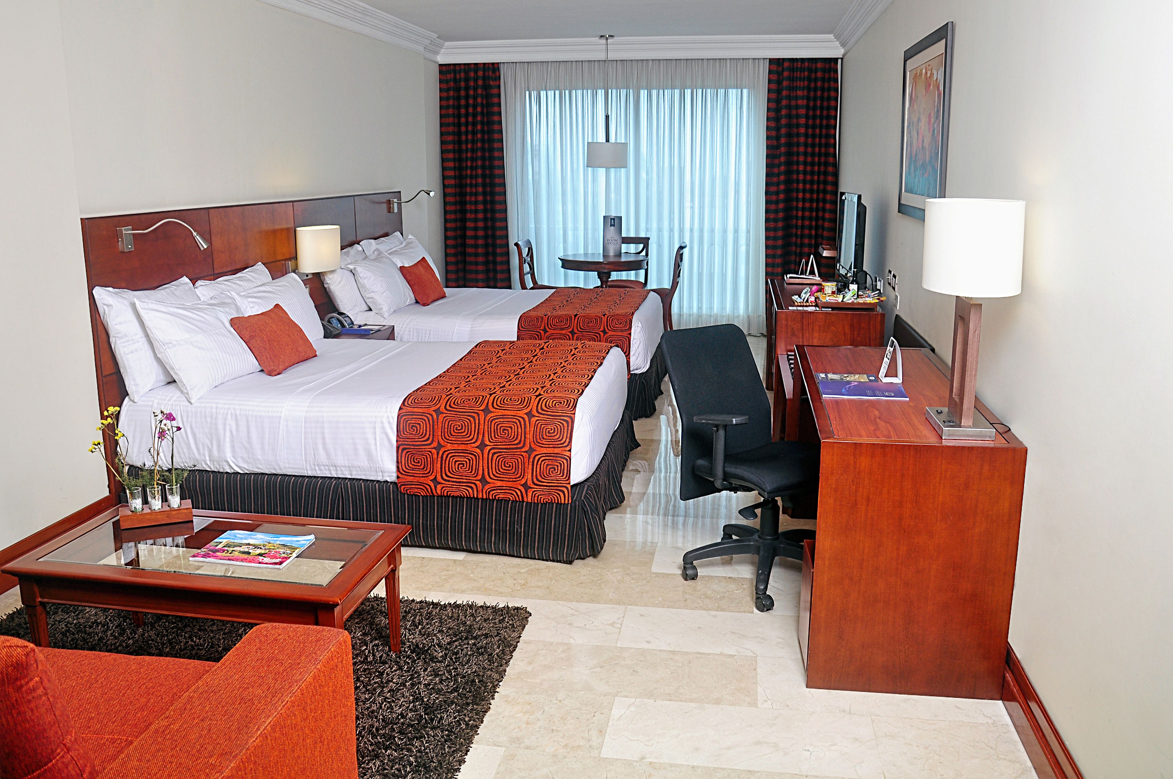 A hotel room with two beds, a desk, a chair, a coffee table, and a TV.