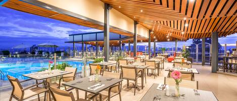 Un área de comedor exterior moderna con mesas y sillas, una piscina al fondo y una estructura de techo de madera.