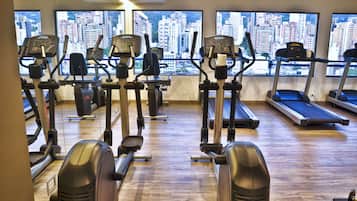 A gym with various cardio machines and a city view through large windows.