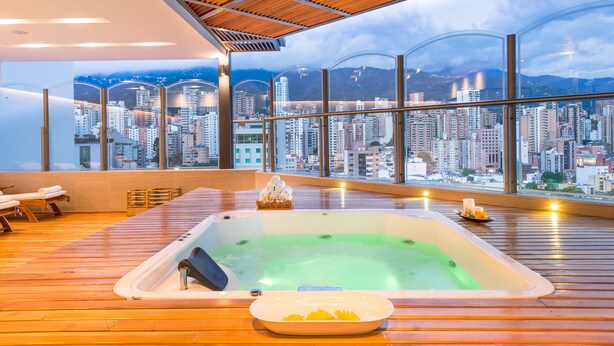 A modern spa area with a large hot tub, wooden flooring, and a panoramic city view through large windows. - Hotel Dann Carlton Bucaramanga (Bucaramanga)